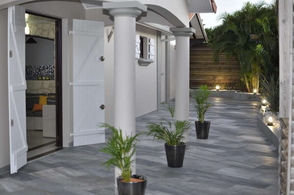 A covered outdoor area with a tiled floor, white columns, and potted plants.