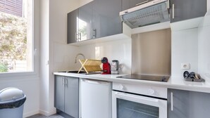 A compact kitchen with grey cabinets, a white countertop, a built-in oven, and a window providing natural light. - Charming apartment located 10 min from the seaside (Marseille)