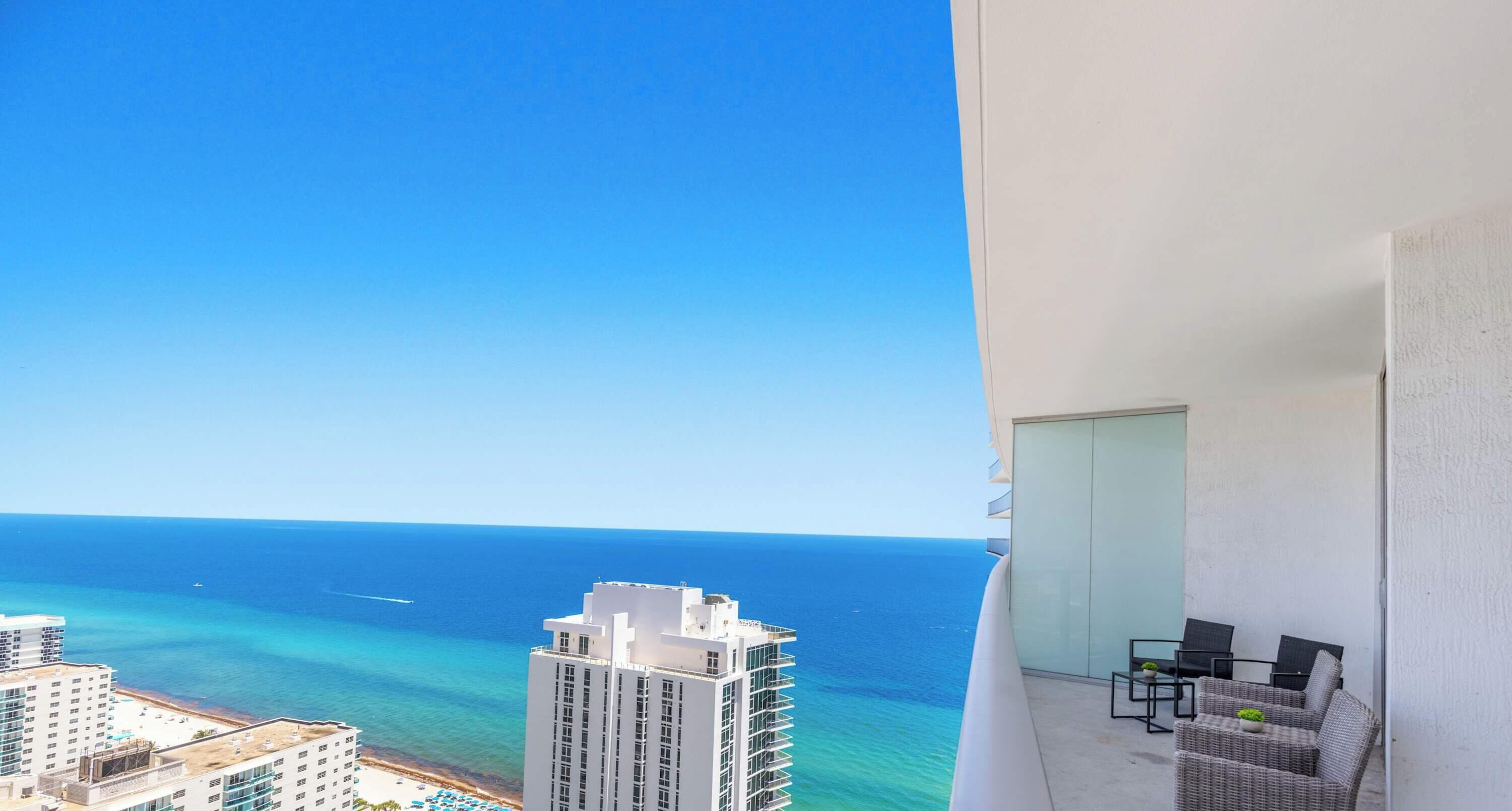 Un balcón con vista al océano y la playa, un área de estar con sillas y una mesa, y un edificio alto al fondo.