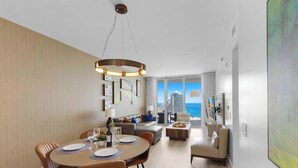 Una habitación de hotel moderna con comedor, un conjunto de sofás, televisor y un ventanal con vista a la ciudad.