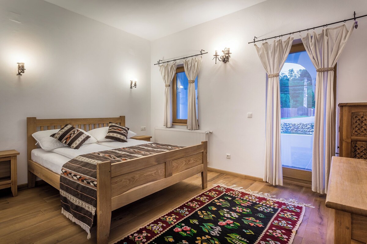 Un dormitorio con cama de madera, mesita de noche, ventana con cortinas, radiador y vistas al exterior.
