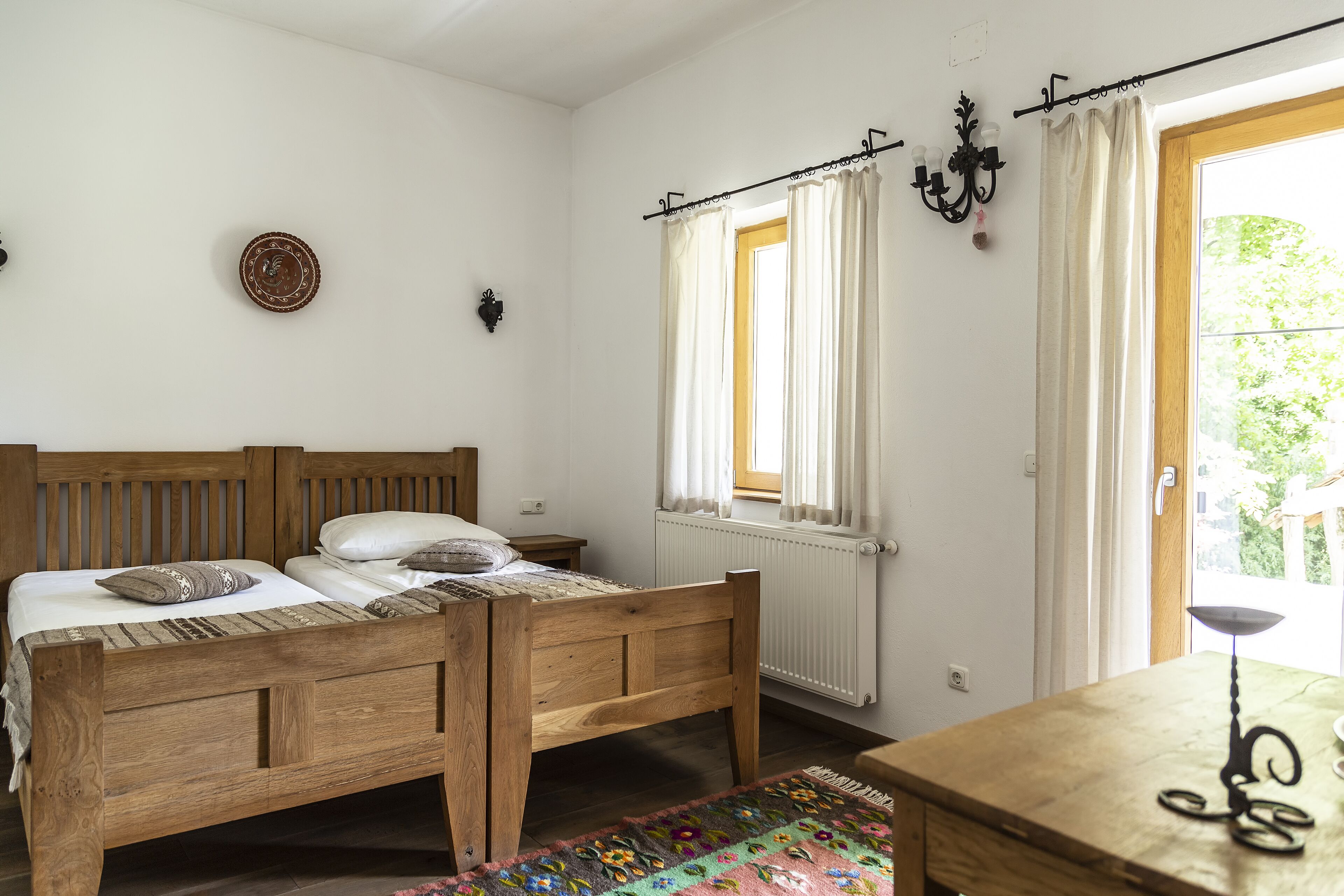 A bedroom with a wooden bed, a radiator, a window with curtains, and a small table with a lamp.