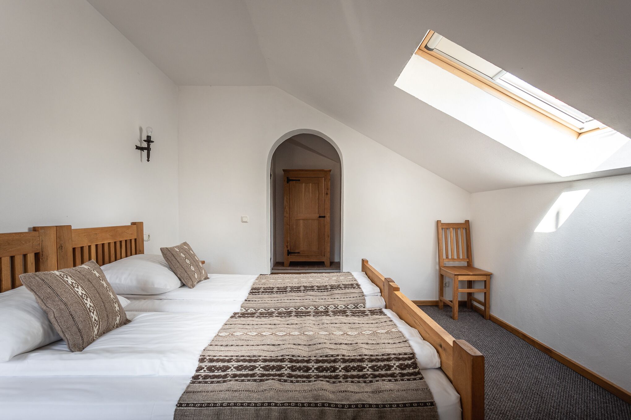 A bedroom with a sloped ceiling, a skylight, a wooden bed, a patterned rug, and a chair.