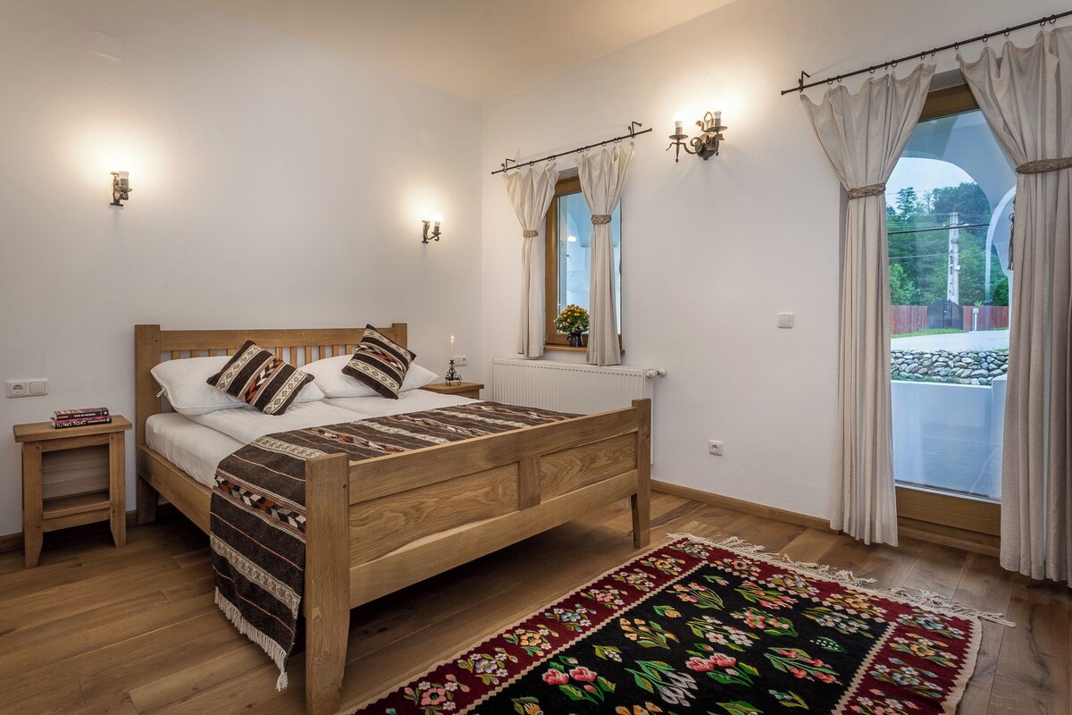 Un dormitorio con cama de madera, mesitas de noche, radiador, ventana con cortinas y vistas al exterior.