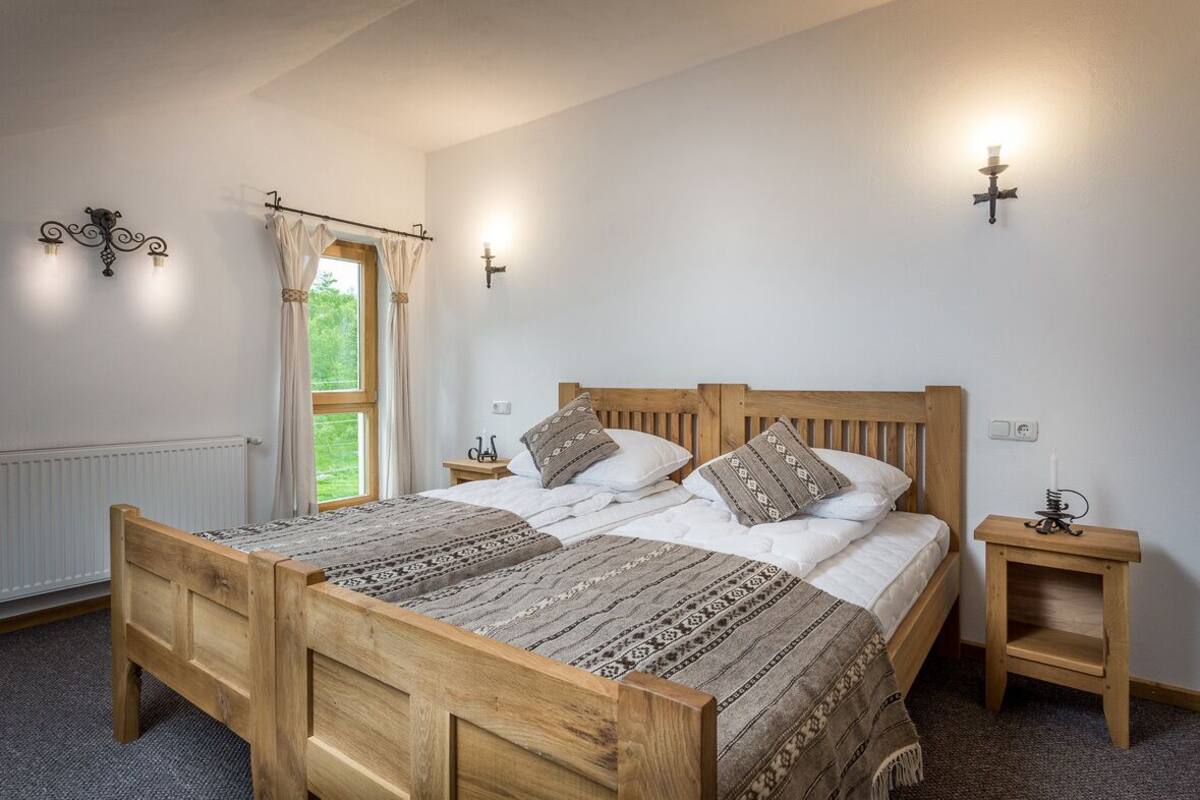A double wooden bed with a patterned bedspread, two bedside tables, a window with curtains, and wall-mounted lighting.