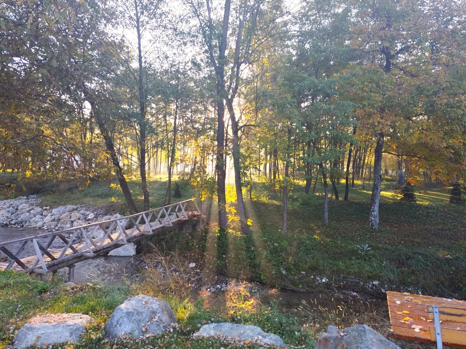 Un pont de fusta sobre un petit torrent en una zona boscosa amb pedres i una banca.