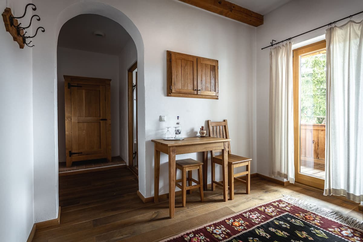 A wooden dining table with chairs, a wooden cabinet, and a rug in a room with wooden flooring and a large window with curtains.