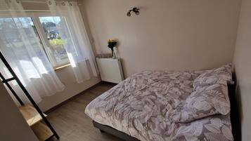 A bedroom with a bed, a radiator, a window with curtains, and a wall-mounted light fixture.