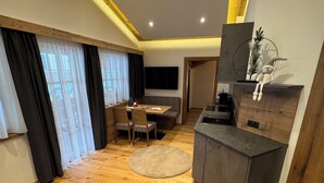 Un saló modern amb sòl de fusta, un sofà gris, una taula de menjador amb cadires, un televisor muntat a la paret i una zona de cuina amb taulell i armaris.