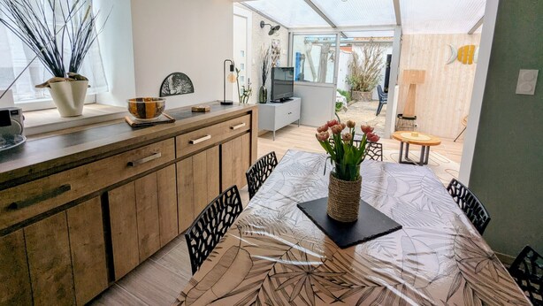 Une cuisine moderne avec des meubles en bois, une table à manger recouverte d’une nappe à motifs et une vue sur le jardin à travers de grandes portes vitrées.
