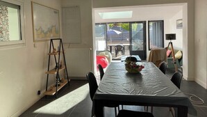 Un coin repas comprenant une longue table, des chaises, un radiateur, une étagère avec des objets décoratifs et une fenêtre avec des stores.
