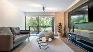 A modern living room with a sofa, a round coffee table, a bean bag, a TV, and a balcony with a view of trees. - w* | Refined 2BR w/ Balcony in Parque España (Ciudad de México)