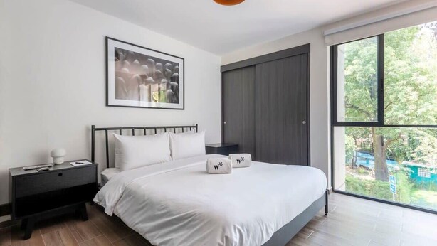 A modern bedroom with a large bed, a nightstand, a framed picture on the wall, and a window with a view of greenery. - w* | Refined 2BR w/ Balcony in Parque España (Ciudad de México)
