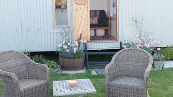 A small, rustic cottage with a wooden porch, two wicker chairs, a small table, and potted plants.