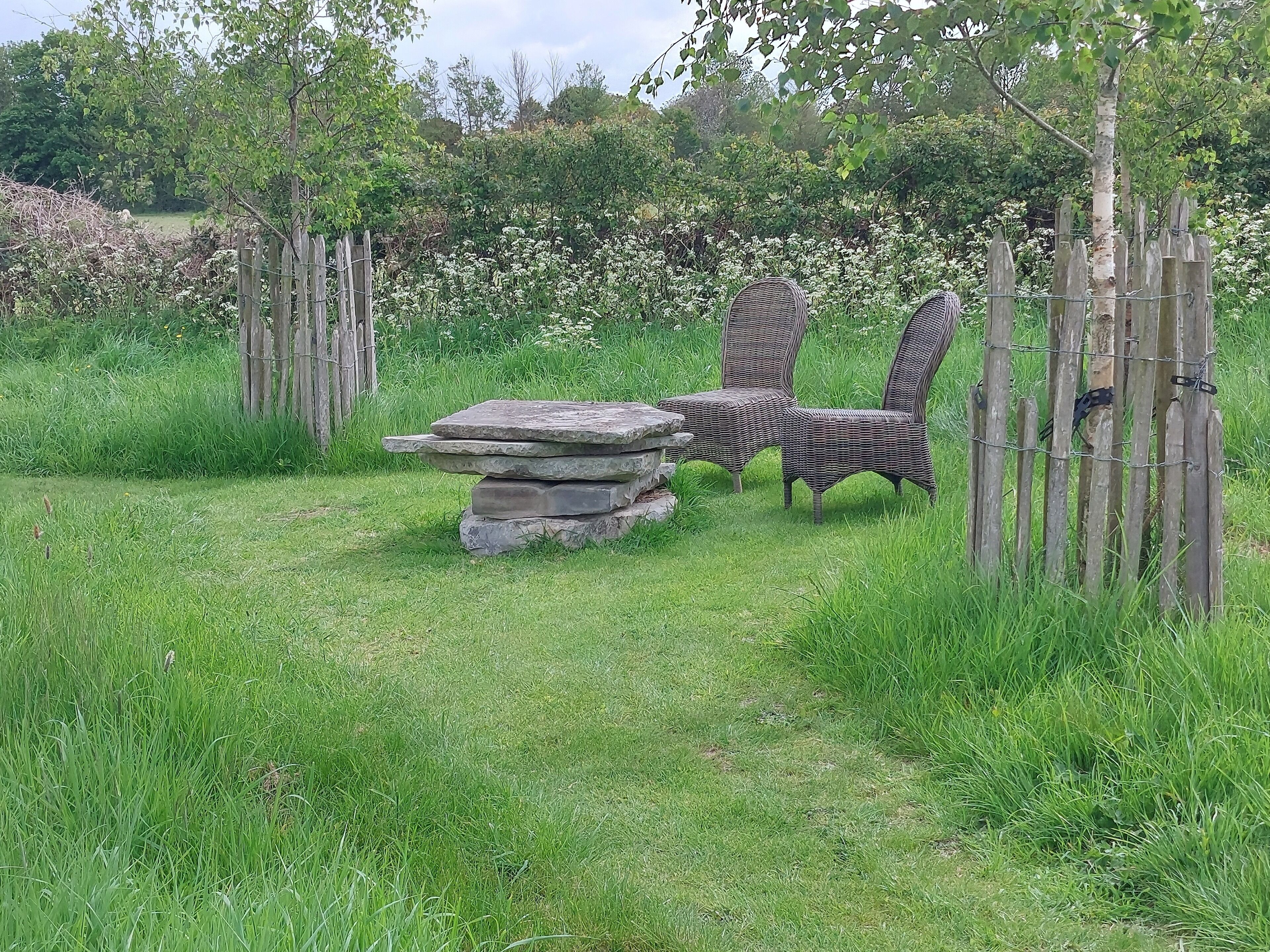 A rustic outdoor seating area with wicker chairs and a stone table, surrounded by a wooden fence and lush greenery.