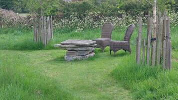 A rustic outdoor seating area with wicker chairs and a stone table, surrounded by a wooden fence and lush greenery.