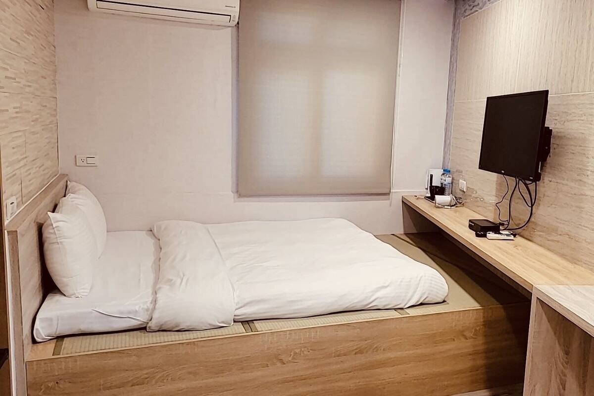 Ein kleines, ordentlich eingerichtetes Schlafzimmer mit einem Bett, einem Holztisch, einem Flachbildfernseher und einem Fenster mit Jalousien.