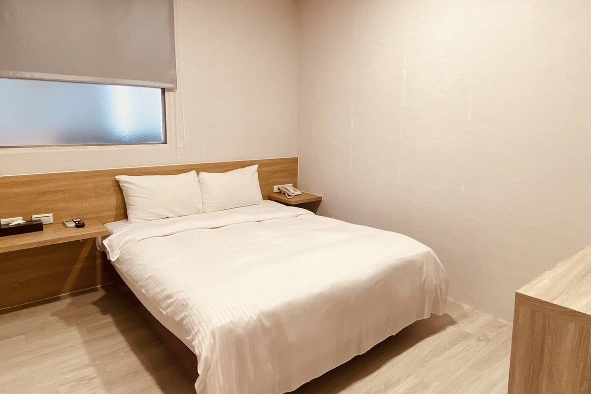 Ein Hotelzimmer mit einem Einzelbett, einem hölzernen Kopfteil, einem Nachttisch mit Telefon und einem Fenster mit Jalousie.