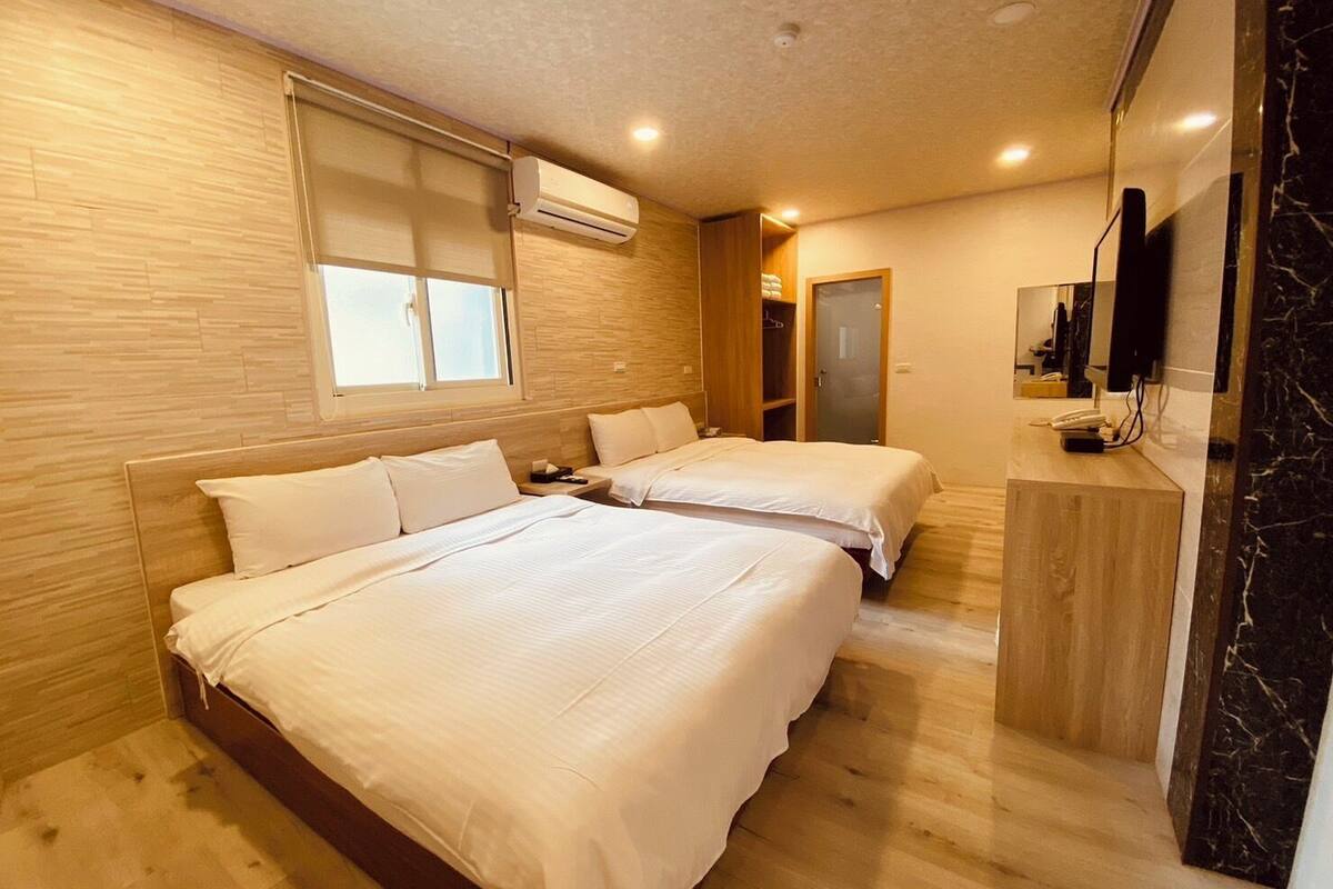Ein Hotelzimmer mit zwei Betten, Holzboden, einem Fenster mit Jalousien, einer Klimaanlage, einem kleinen Schreibtisch mit Telefon und einem Spiegel an der Wand.