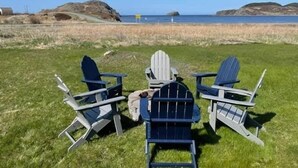 Three Adirondack chairs facing a grassy area with a view of the ocean and distant hills. - Red Sunset Cottage in Twillingate (Twillingate)