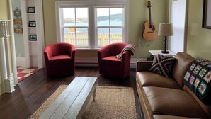 Un salon avec un canapé en cuir marron, deux fauteuils rouges, une guitare et une vue sur la mer grâce à de grandes fenêtres.