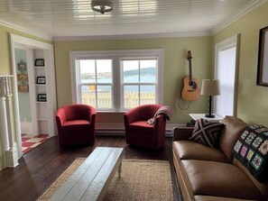 A living room with a brown leather sofa, two red armchairs, a guitar, and a view of the sea through large windows.