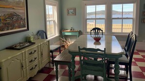 Une salle à manger avec un sol à carreaux, une table entourée de chaises, une fenêtre donnant sur la mer et un tableau accroché au mur.