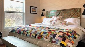 A bedroom with a large bed, a wooden headboard, a window, and a bench. - Spencer Salt-Side Cottage (Salt Harbour) (Herring Neck)