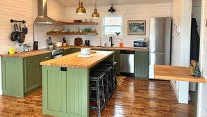 A kitchen with wooden cabinets, a central island, bar stools, a stainless steel refrigerator, and hanging pots. - Spencer Salt-Side Cottage (Salt Harbour) (Herring Neck)