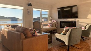 A living room with a sofa, armchairs, a fireplace, a TV, and large windows overlooking a body of water. - Spencer Salt-Side Cottage (Salt Harbour) (Herring Neck)