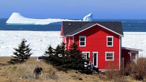 A red house with a small porch, a clear view of the ocean, and an iceberg in the background. - Twilly House (Twillingate)