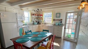 A kitchen with wooden floors, a dining table with colorful chairs, a refrigerator, and a window with curtains. - Twilly House (Twillingate)