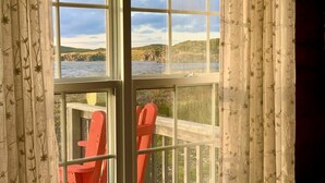 A room with a large window overlooking a body of water and a wooden deck with red chairs. - Twilly House (Twillingate)