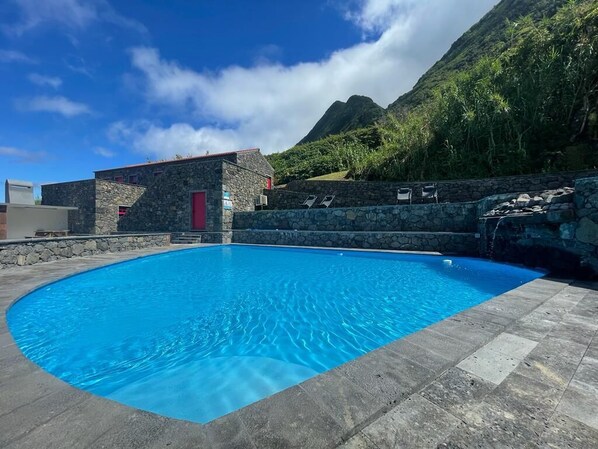 A swimming pool with a stone wall and a red door, surrounded by a mountainous landscape.