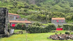 A stone building with red doors and a red roof, surrounded by lush greenery and a hillside. - Stylish 1 Bedroom Stone House With Sea View In São Jorge, Azores (Belo Malandro) (São Jorge)
