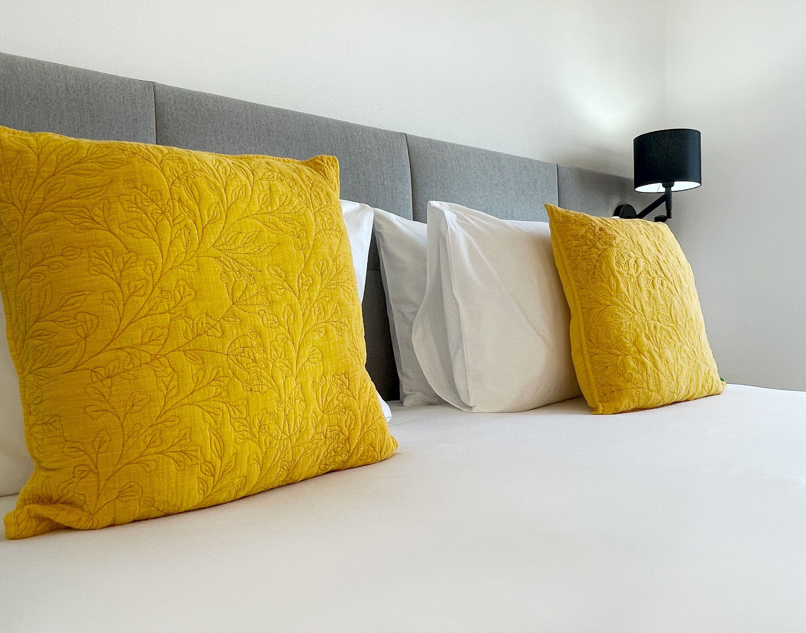 Letto con tre cuscini, uno grande giallo e due più piccoli bianchi, appoggiati a una testiera grigia.