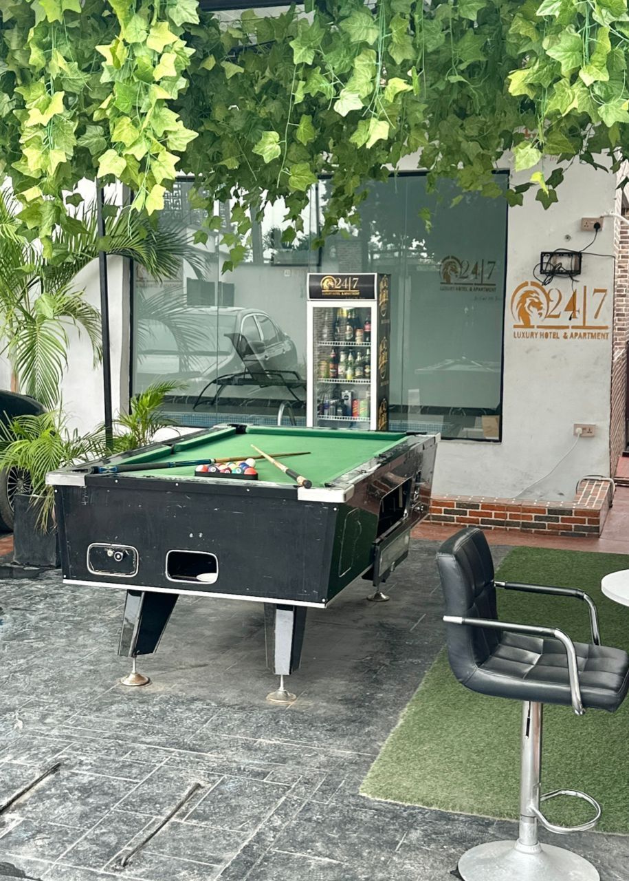 A pool table with a green felt surface, a pool cue, and several balls. There is a bar area with a black chair and a bar stool. The setting appears to be outdoors with a building in the background.