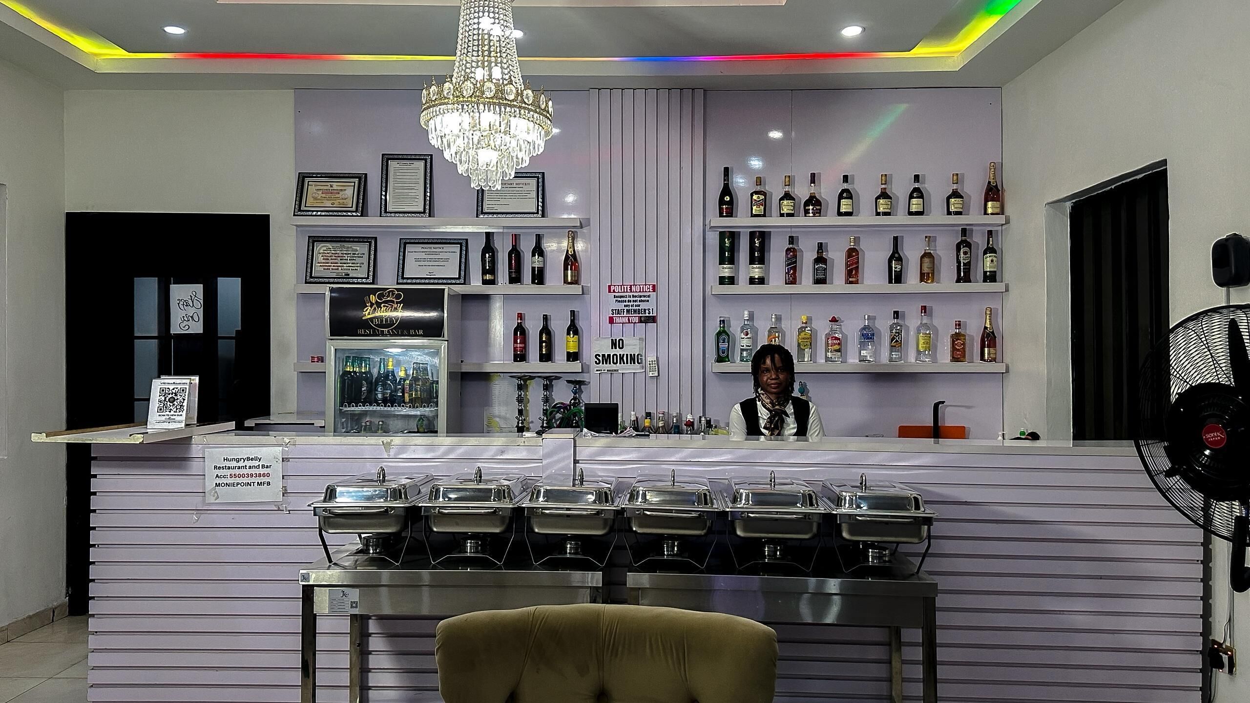 A bar area with a counter, shelves of liquor bottles, a chandelier, and a 'No Smoking' sign.