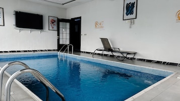 An indoor swimming pool with clear blue water, a metal ladder, and a lounge chair.