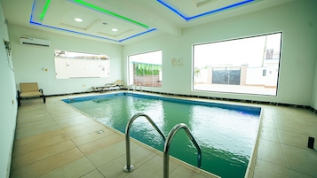 An indoor swimming pool with clear blue water, a metal ladder, and a view of a residential area through large windows.