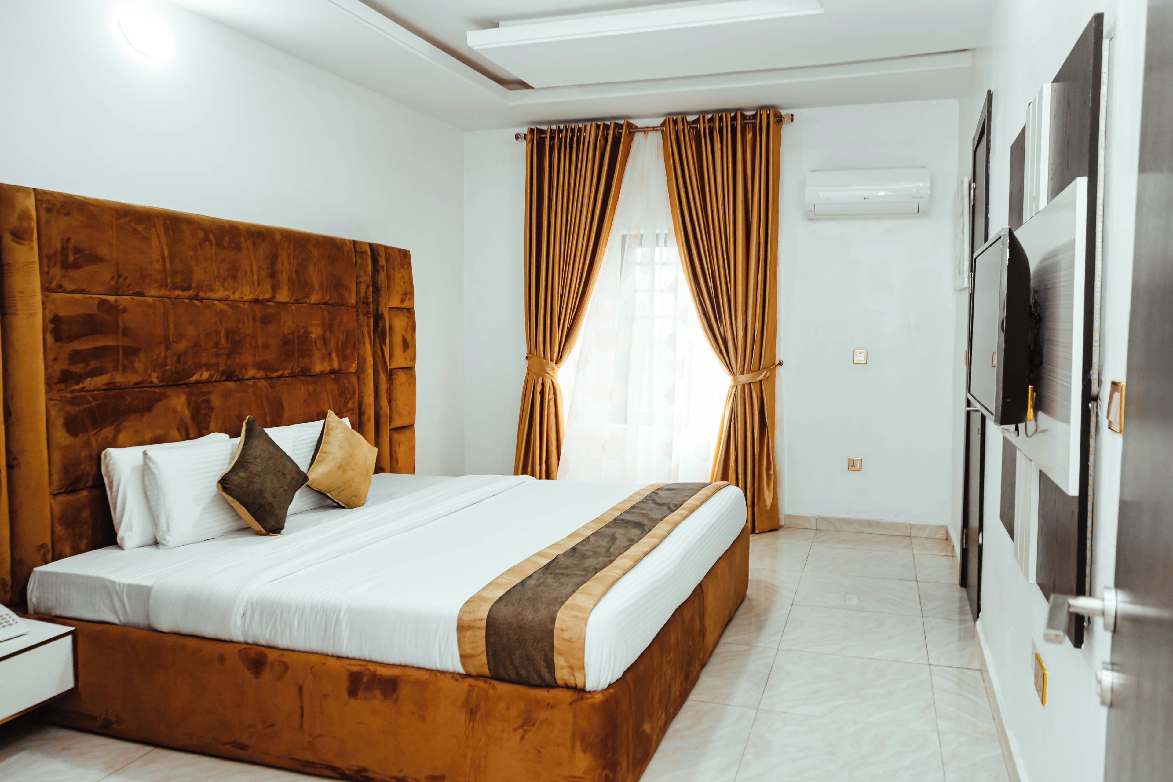 A hotel room with a large bed, a wooden headboard, a nightstand, a TV, and a window with curtains.