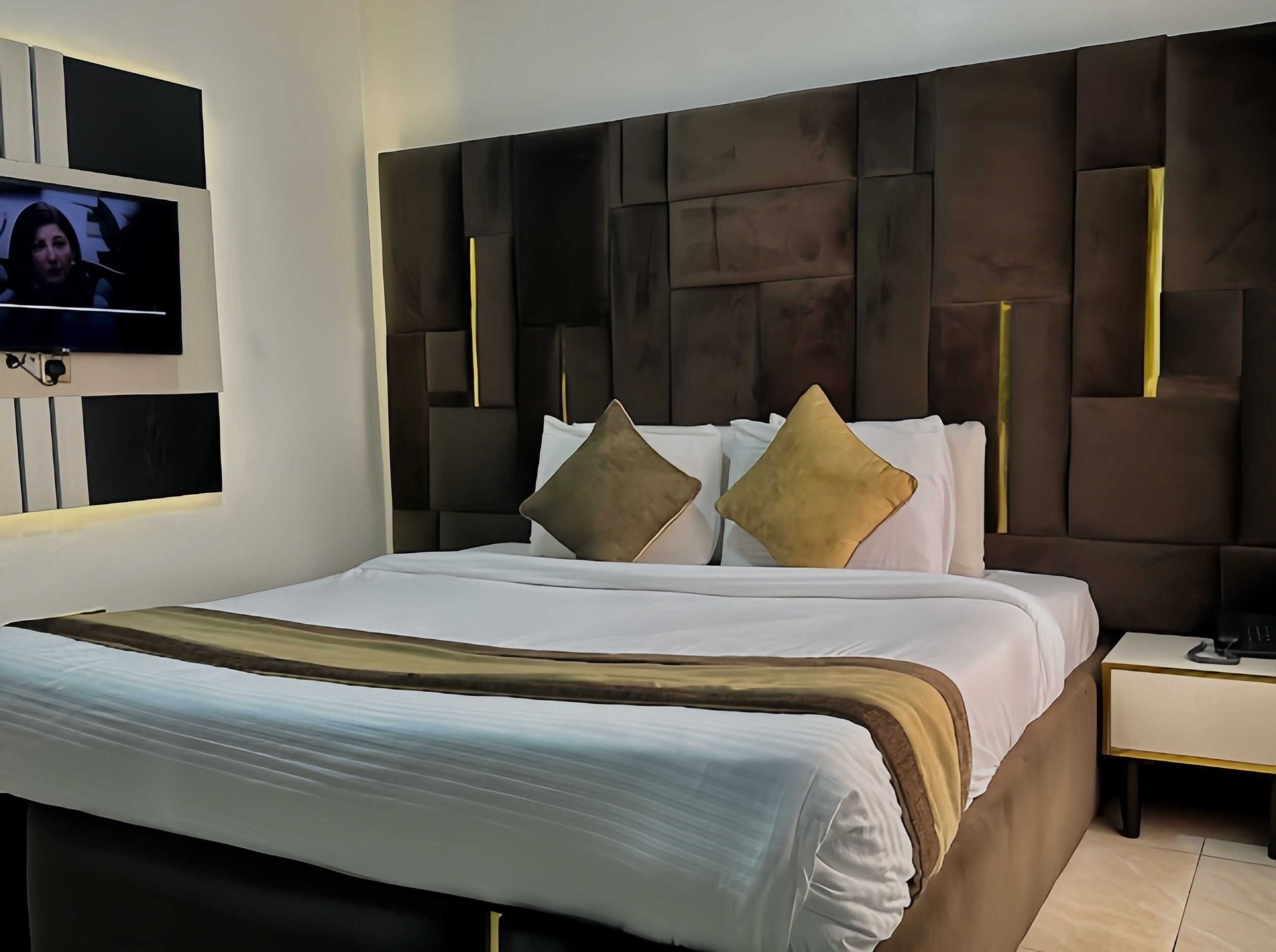 A modern hotel room with a large bed, a flat-screen TV mounted on the wall, and a nightstand with a lamp.