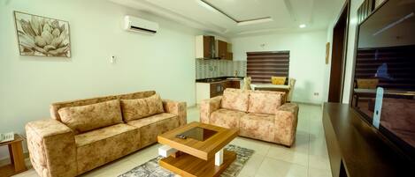 A living room with a sofa, a coffee table, and a TV. There is a kitchen in the background.