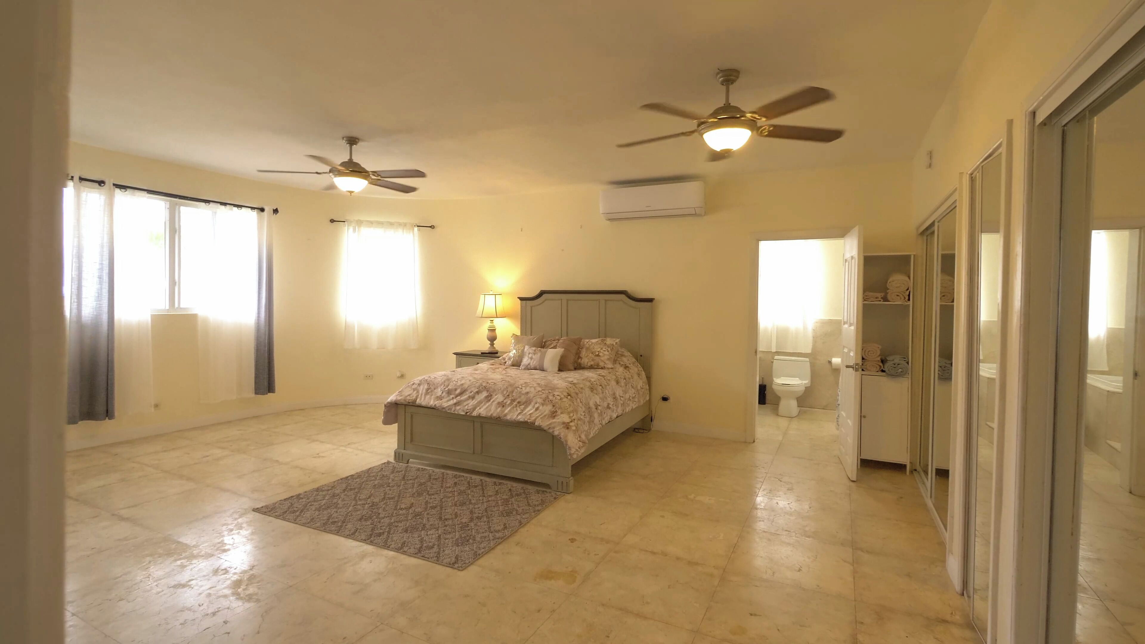 A bedroom with a bed, two ceiling fans, a nightstand, a lamp, a bathroom with a toilet and a shower, and a walk-in closet.