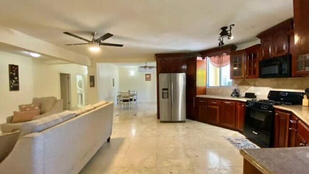 A spacious living room with a sofa and a dining area in the background. A kitchen with wooden cabinets and a refrigerator is visible.