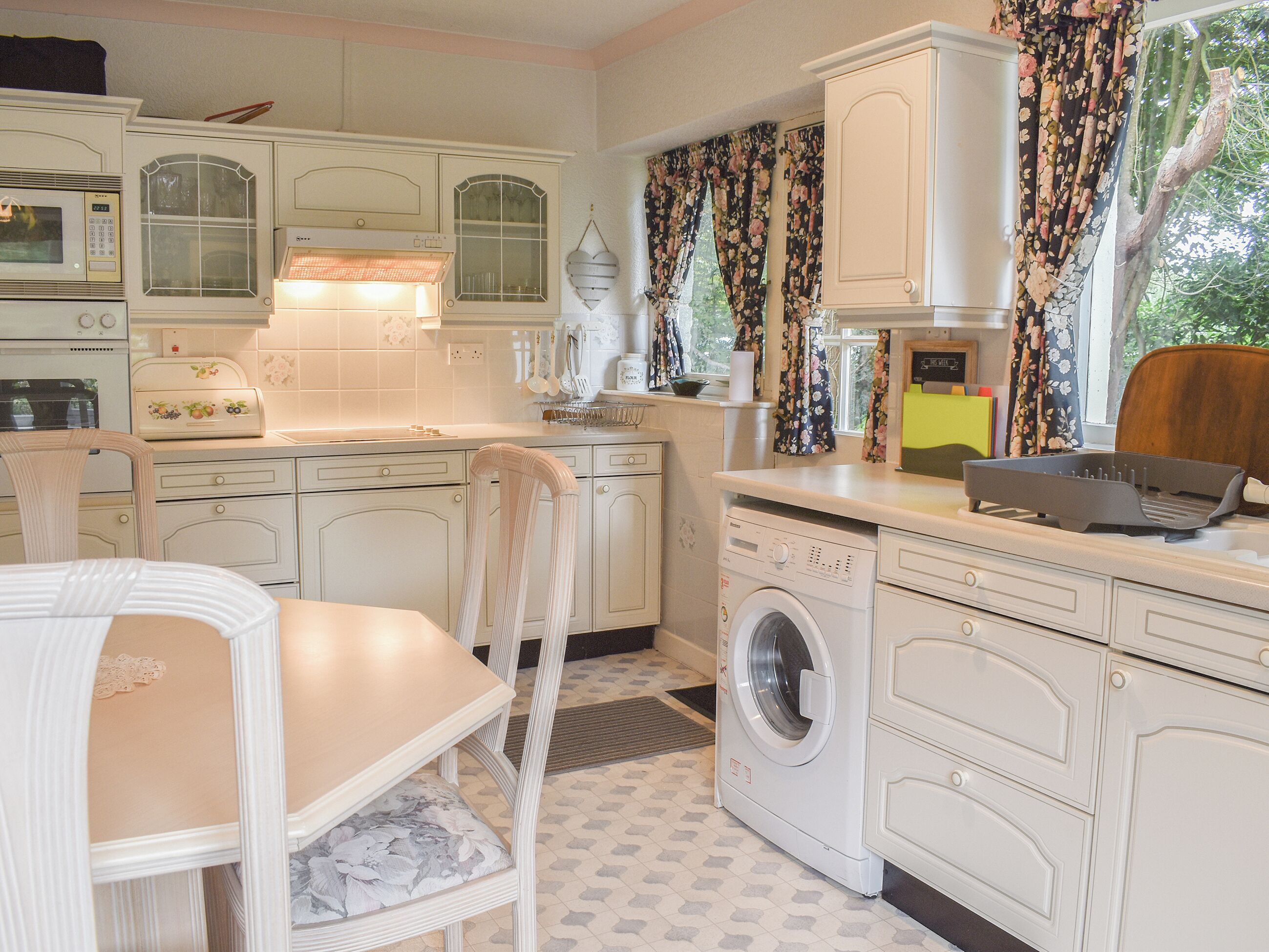 Een kleine keuken met witte kasten, een wasmachine en een eethoek met een stoel.