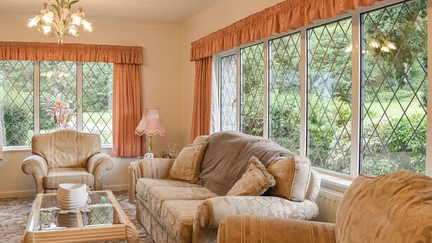 A room with a sofa, armchair, and a glass coffee table. There are large windows with curtains, and a chandelier is visible.