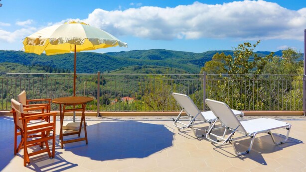 Una terrazza con tavolo e sedie, un ombrellone giallo e bianco e vista su dolci colline verdi.