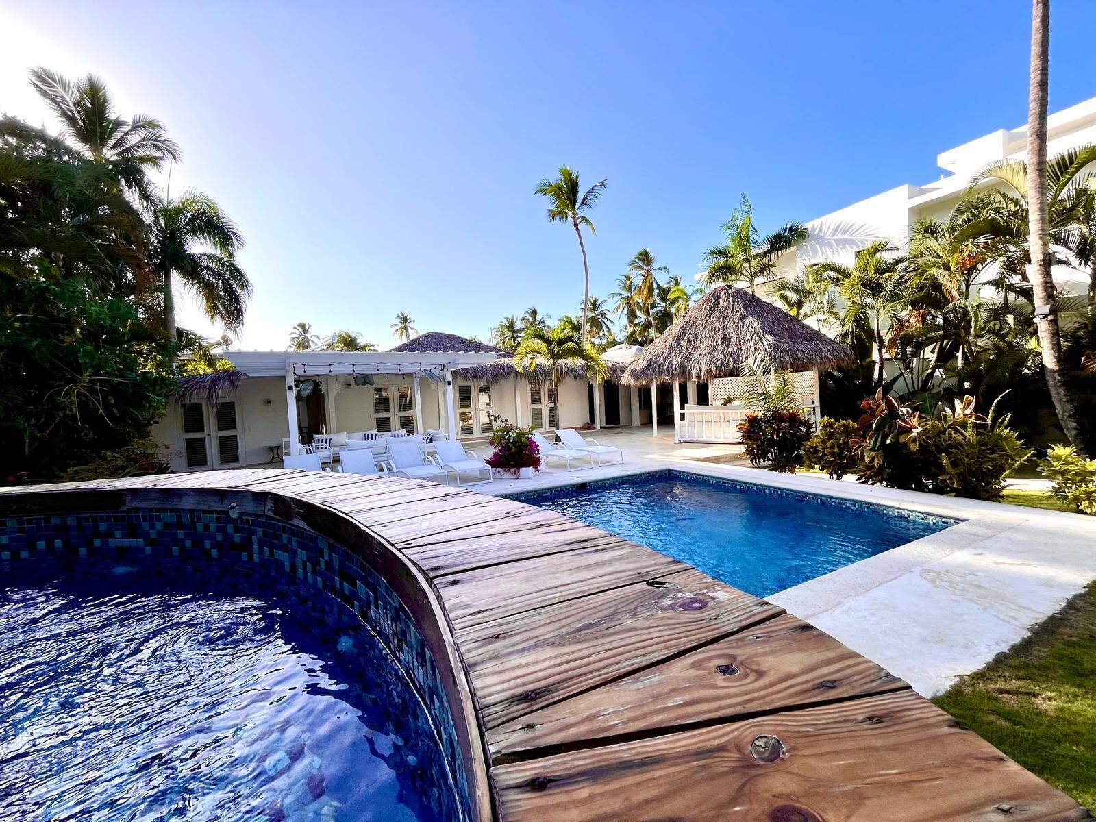 A pool area with a wooden deck, lounge chairs, and a thatched-roof structure.