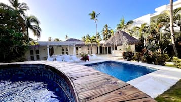 A pool area with a wooden deck, lounge chairs, and a thatched-roof structure.